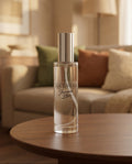 A clear cylindrical perfume bottle with a reflective silver cap stands on a round wooden table in a softly blurred living room. The bottle label reads 'LUCEVERA Japanese Plum'.