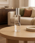 A clear glass perfume bottle with a silver cap stands on a round wooden table in a softly lit living room. The bottle label reads LUCEVERA and appears to be the product being sold.