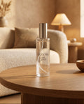 A clear glass bottle of Acevera Black Bean room spray with a metallic silver cap, set on a round wooden table in a warm, softly lit living room.