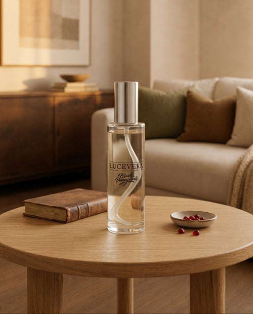 A clear rectangular perfume bottle with a silver cap stands on a round light wood table in a softly lit living room. The bottle label reads 'Lucevera' and 'Black Pomegranate'.